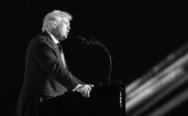 A black-and-white photograph of Donald Trump delivering a speech at a lectern, taken from the side