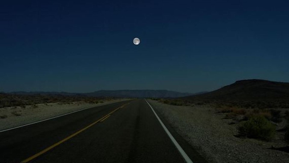 A photograph of a deserted road through a desert valley, with the full moon hanging in the sky.