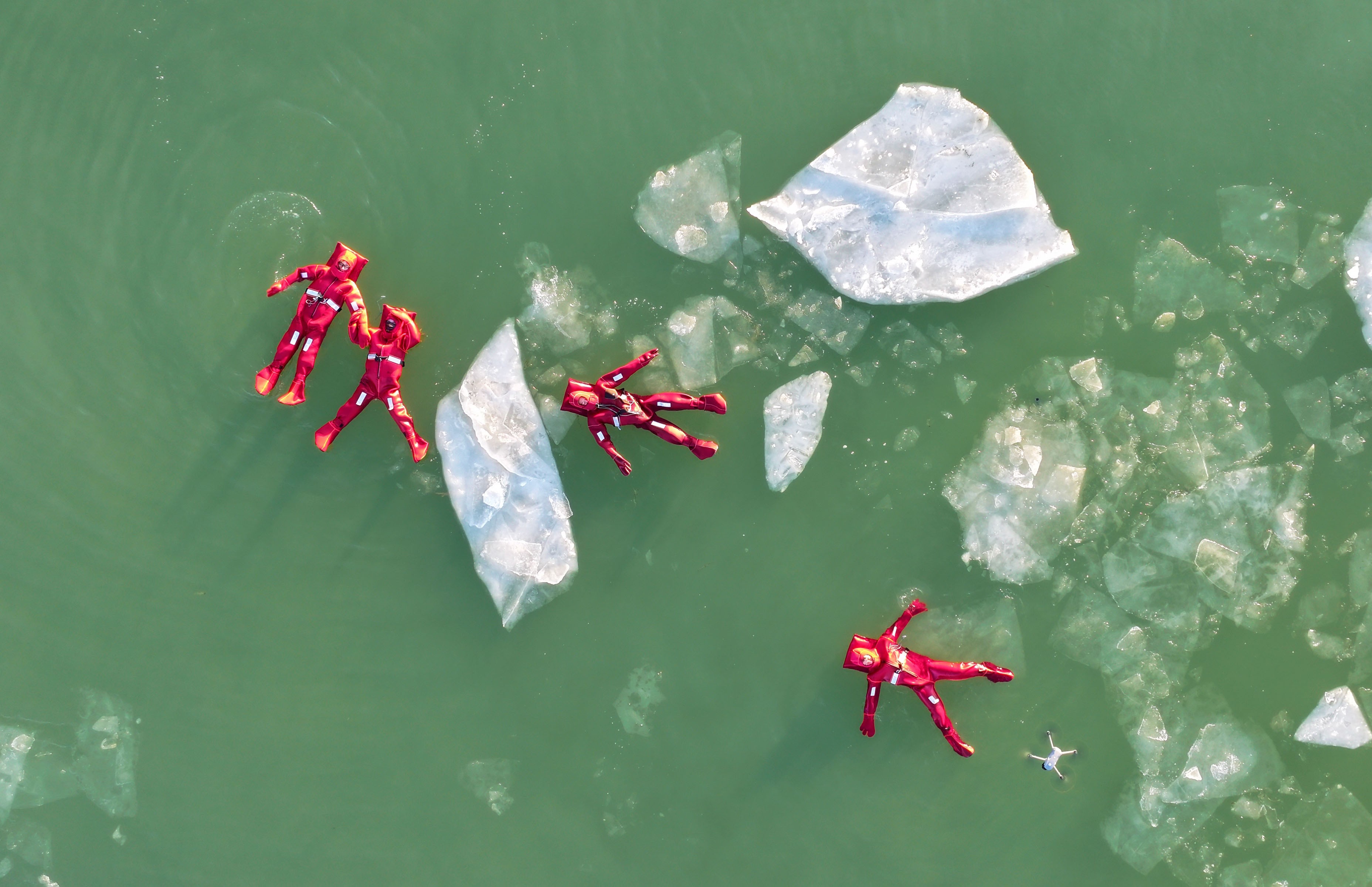 An aerial view of four people wearing bright red rubber protective suits, floating in green-colored water among chunks of floating ice