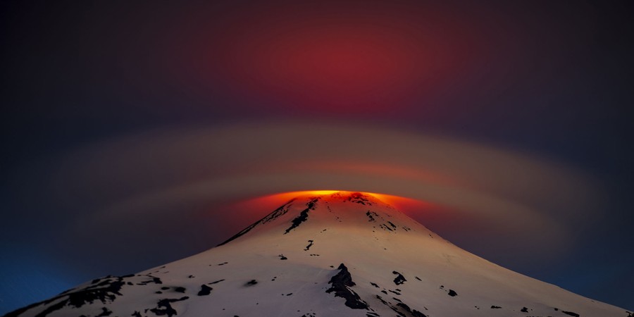 Smooth clouds cap an erupting volcano at night, glowing orange from within.