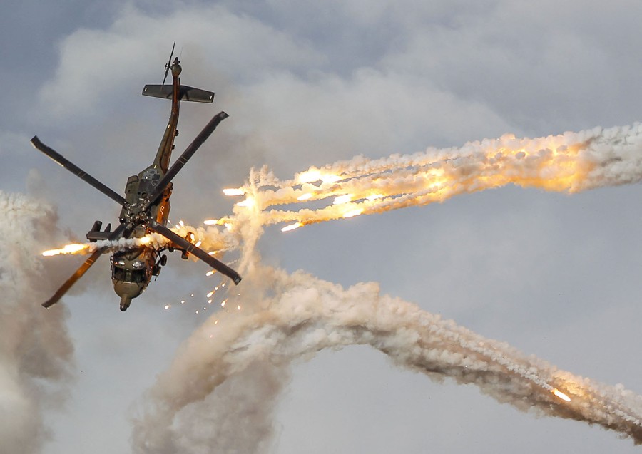 A military helicopter flies at a steep angle past flares and streams of smoke.