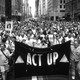 A big group of protesters march down a street holding an ACT UP banner.