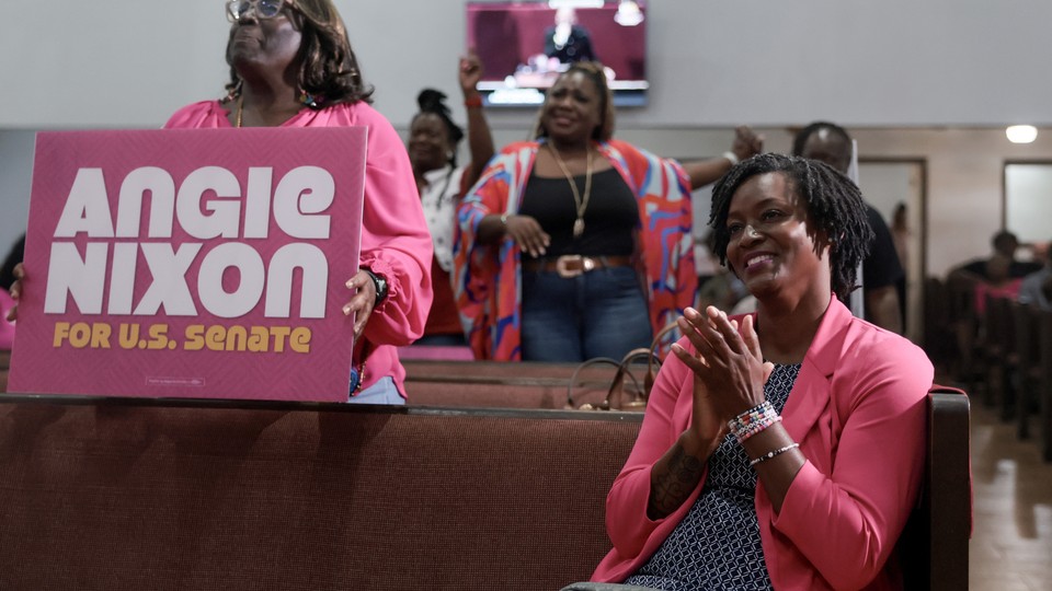 Photograph of Angie Nixon wearing a pink blazer. She is seated and applauding while a woman holding an “Angie Nixon” sign stands near her.
