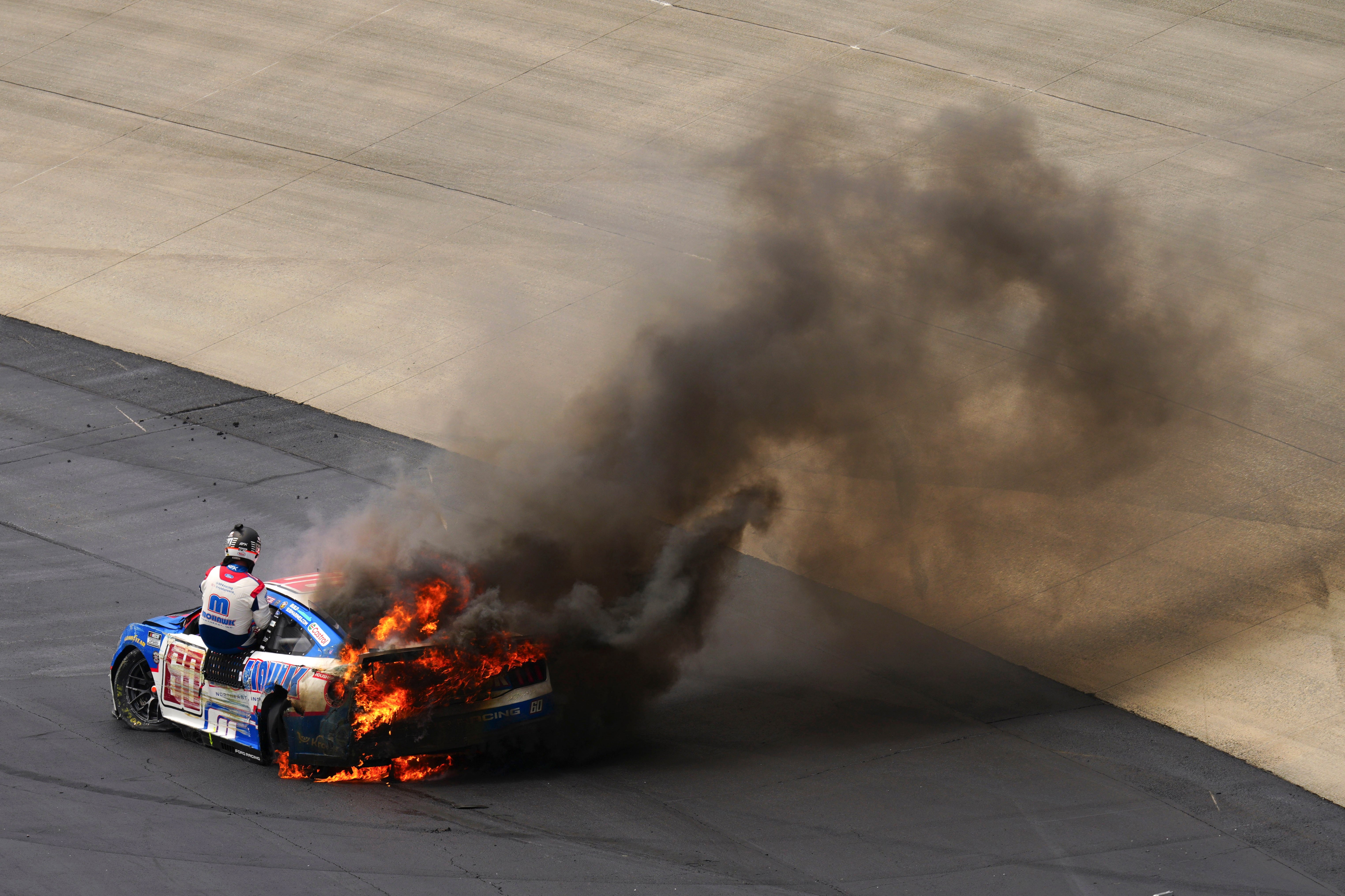 A NASCAR driver climbs out the window of a burning race car.