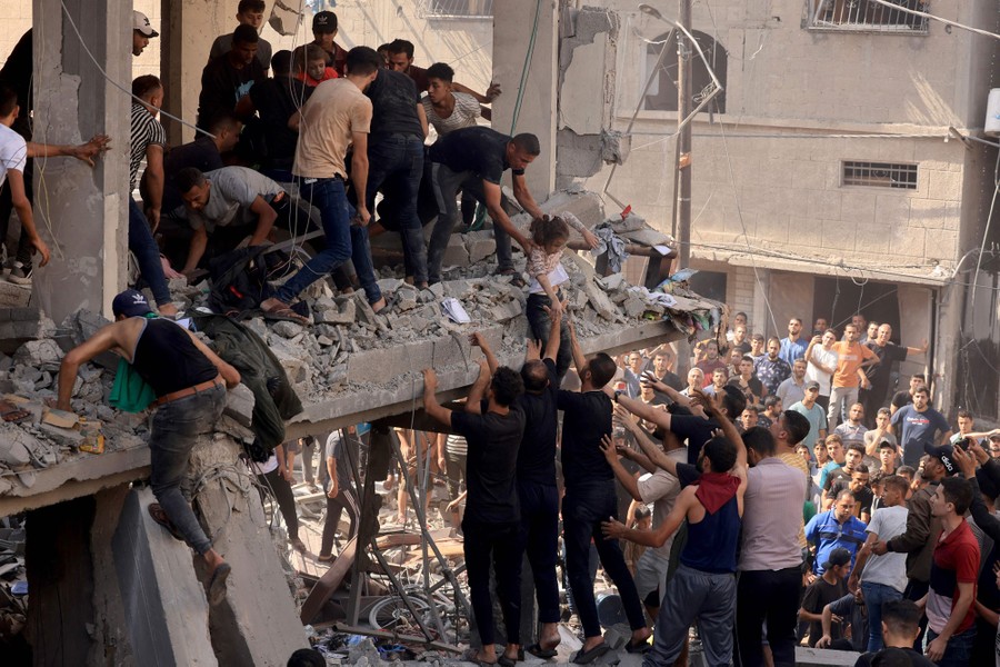 A large crowd of people works to lower a child from the debris-filled second floor of a heavily damaged building.