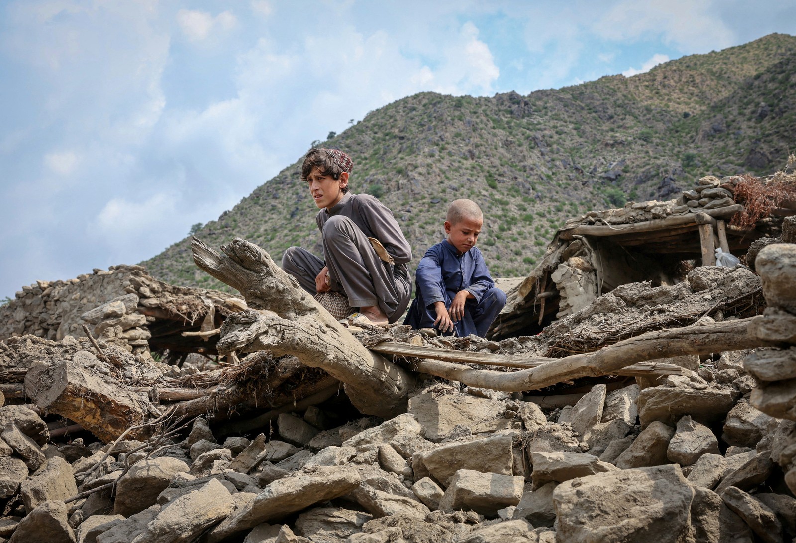 Two boys sit atop a pile of rubble that was once a house made of stone and timbers.