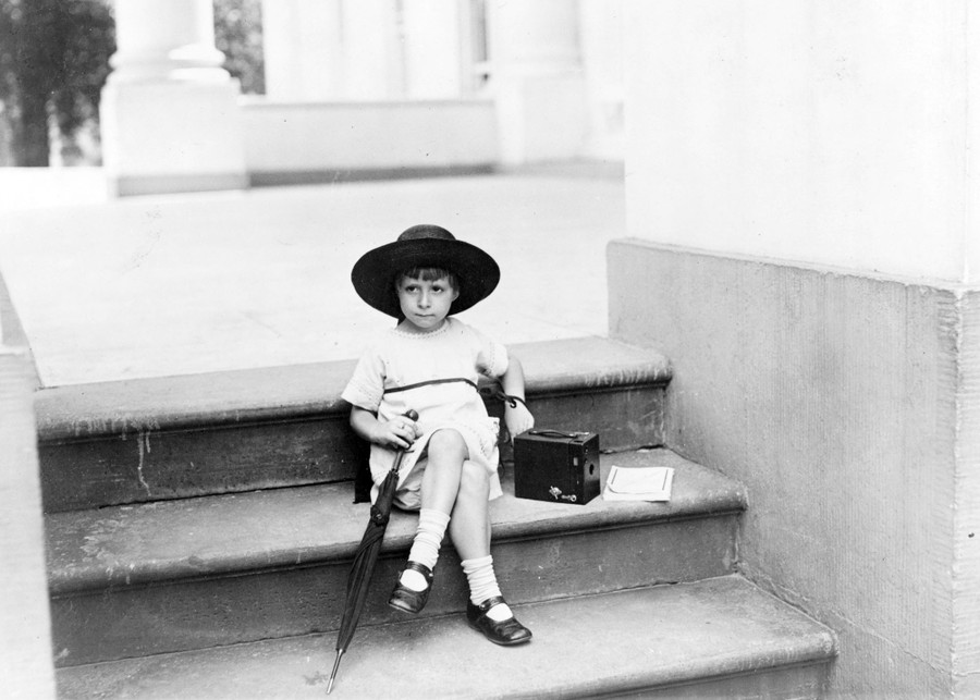 A girl sits on steps outdoors, with an umbrella and a handheld camera.