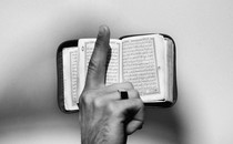 A photograph of a raised finger in front of a Quran