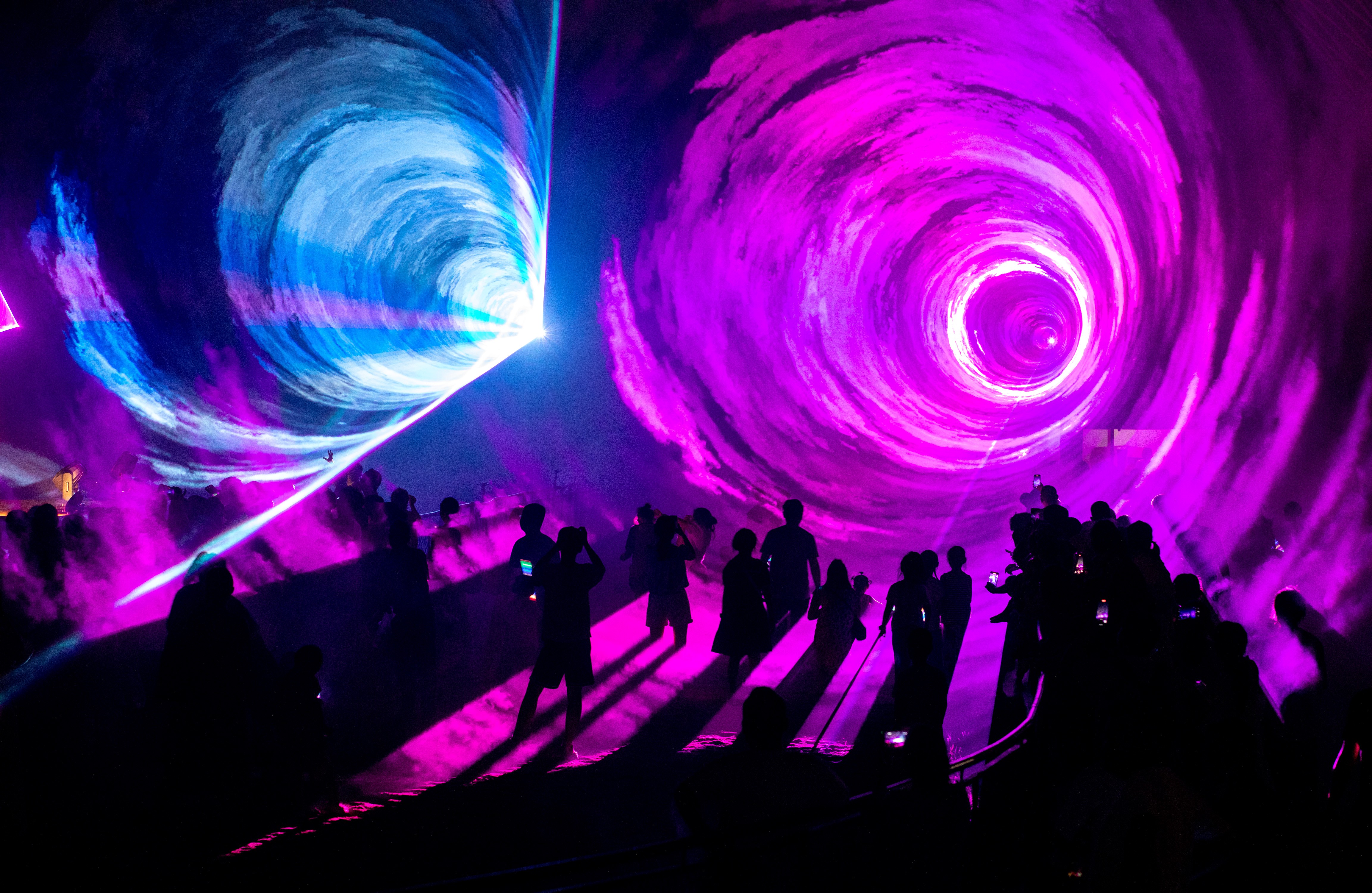 People attend a laser show, standing in colorful tunnels of light.