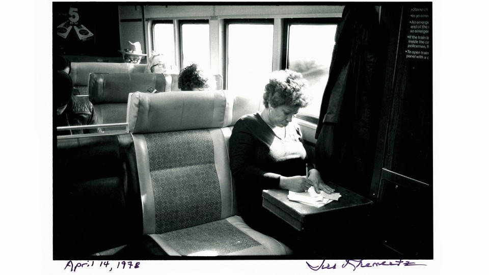 black-and-white photo of woman sitting in window seat on train, head bowed and writing on a briefcase on her lap, signed at bottom by photographer and hand-dated April 14, 1978