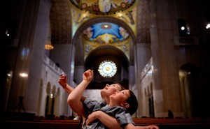 A woman holds a child in her arms as they both look up toward the ornate ceiling of a basilica.