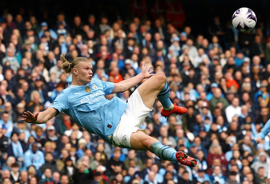 A soccer player jumps while kicking a ball, with a crowd of onlookers in the background.