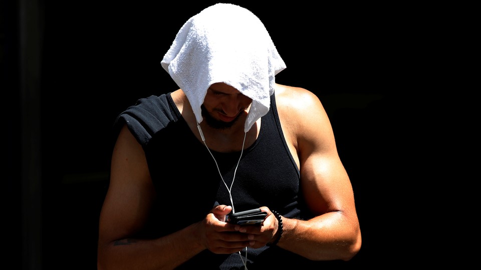 A man wearing a tank top covers his head with a towel and looks down at his phone.
