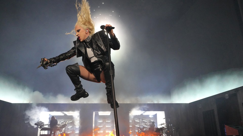 Lady Gaga in midair at her microphone on a stage covered in flames