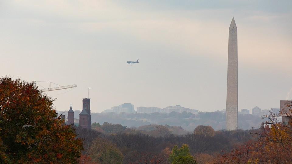 A plane flies in the sky over trees with multicolored leaves as the Washington Monument stands in the background.