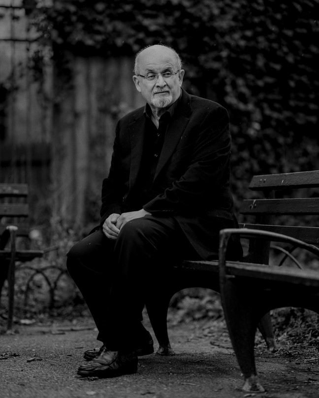 A black-and-white photograph of Salman Rushdie sitting on a park bench