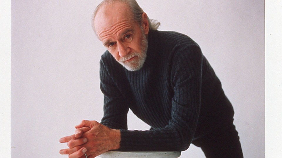 Headshot of George Carlin leaning forward on a bench