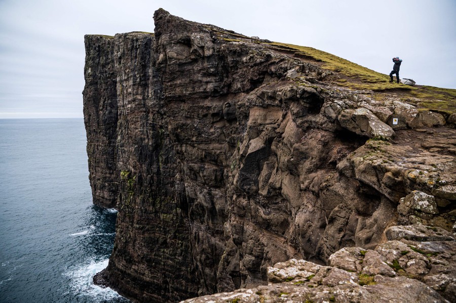 A person stands near a tall, steep sea cliff, looking toward the ocean.