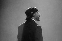 A black-and-white photo of Bad Bunny standing alone onstage in a suit and backward cap