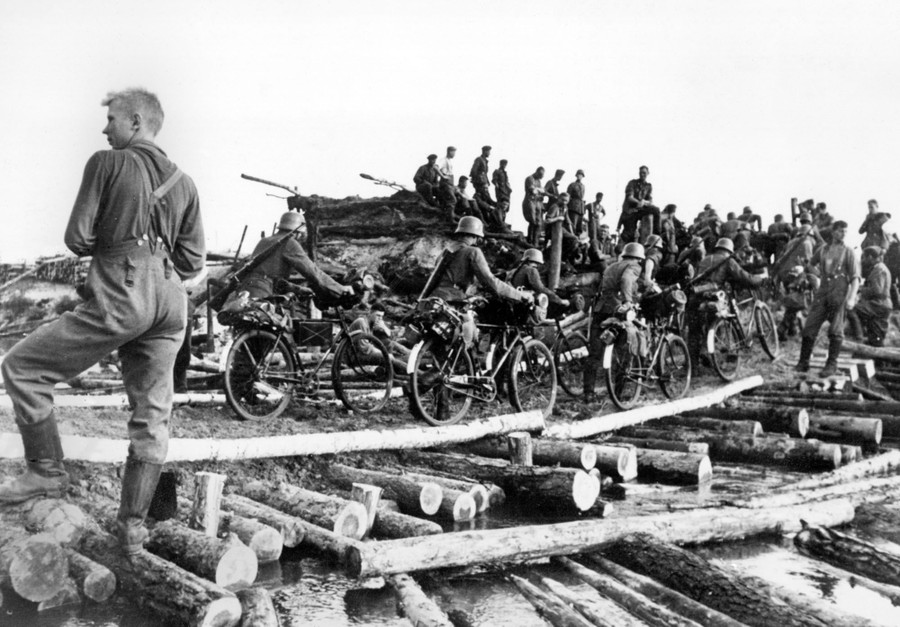 A unit of German cyclists walks over a hastily built roadway made of logs.