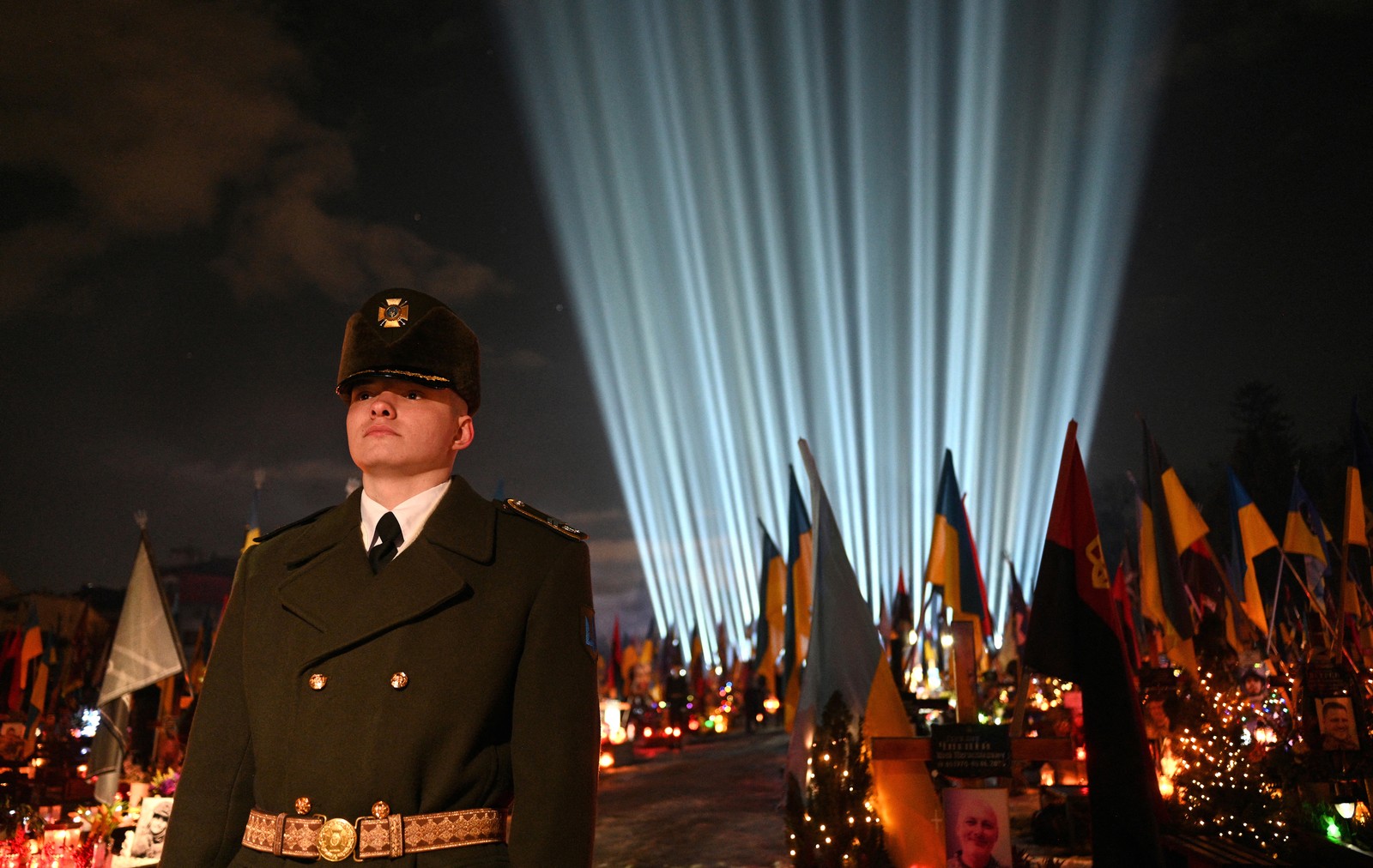 A soldier in dress uniform stands guard in front decorated military gravestones and a light display.