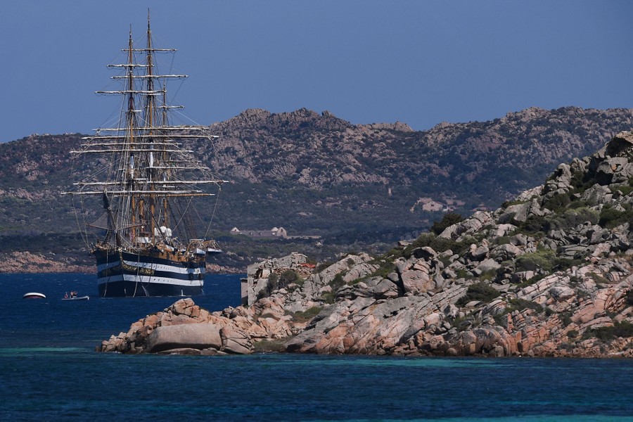 A tall masted ship sits at anchor off the coast of a rocky island.
