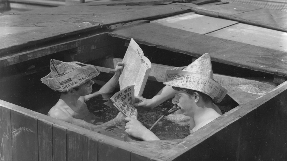 Two boys wearing newspaper boat hats and reading the newspaper in a wooden tub.