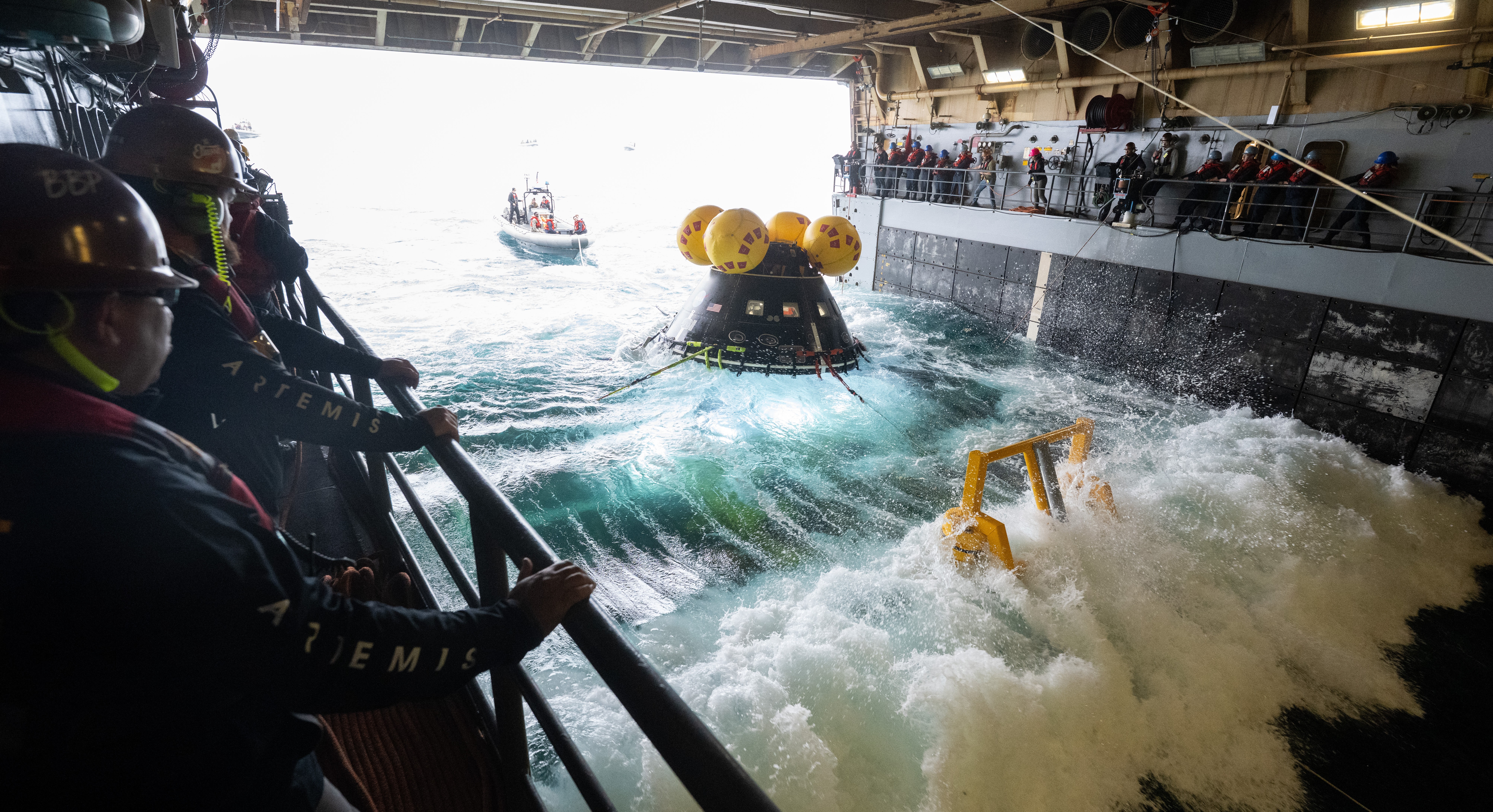 People stand on decks inside an open space at the bottom of a ship, on either side of a space capsule bobbing in waves.