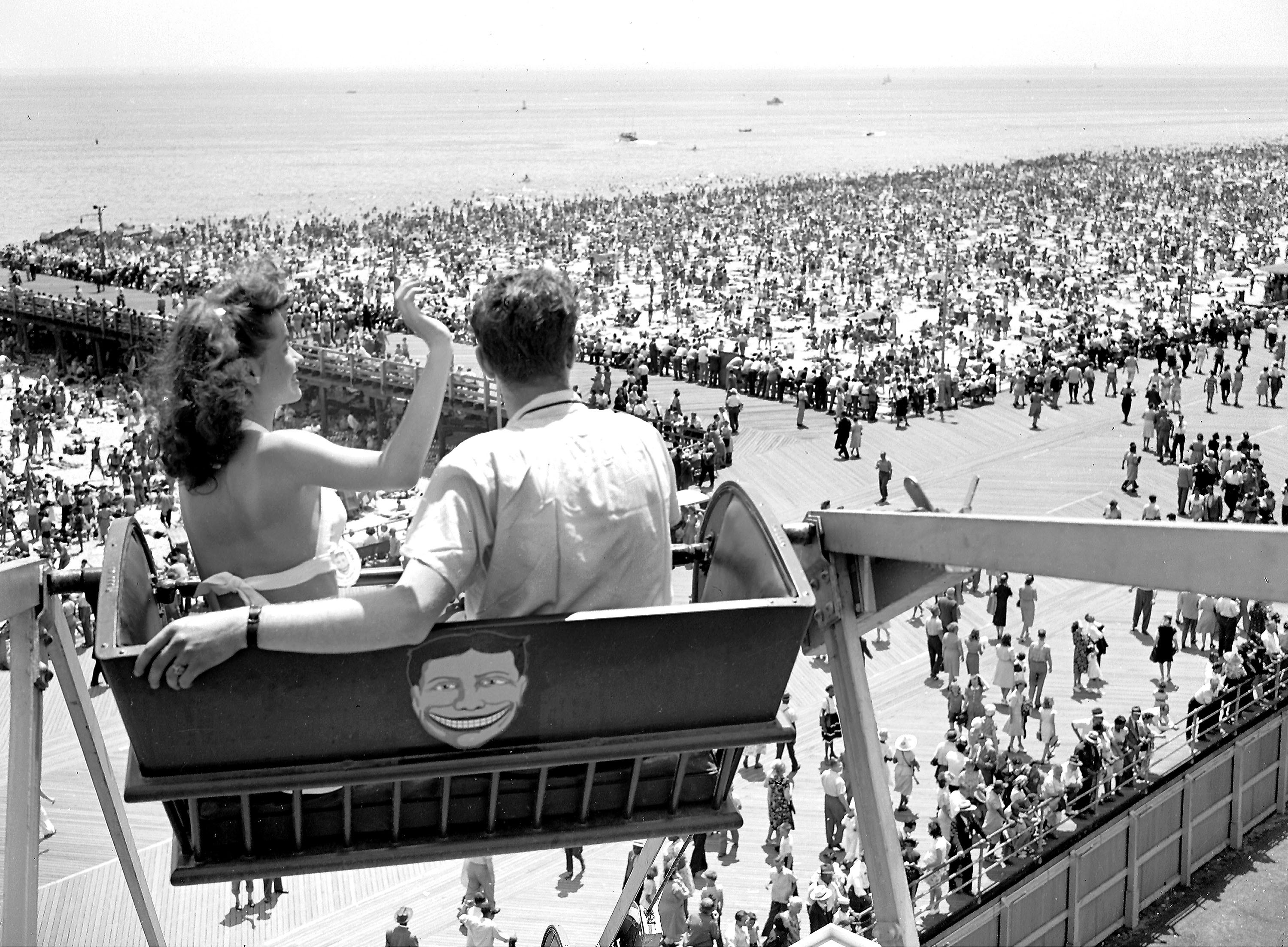 A couple sits high on a Ferris wheel, looking out over a crowded beach.