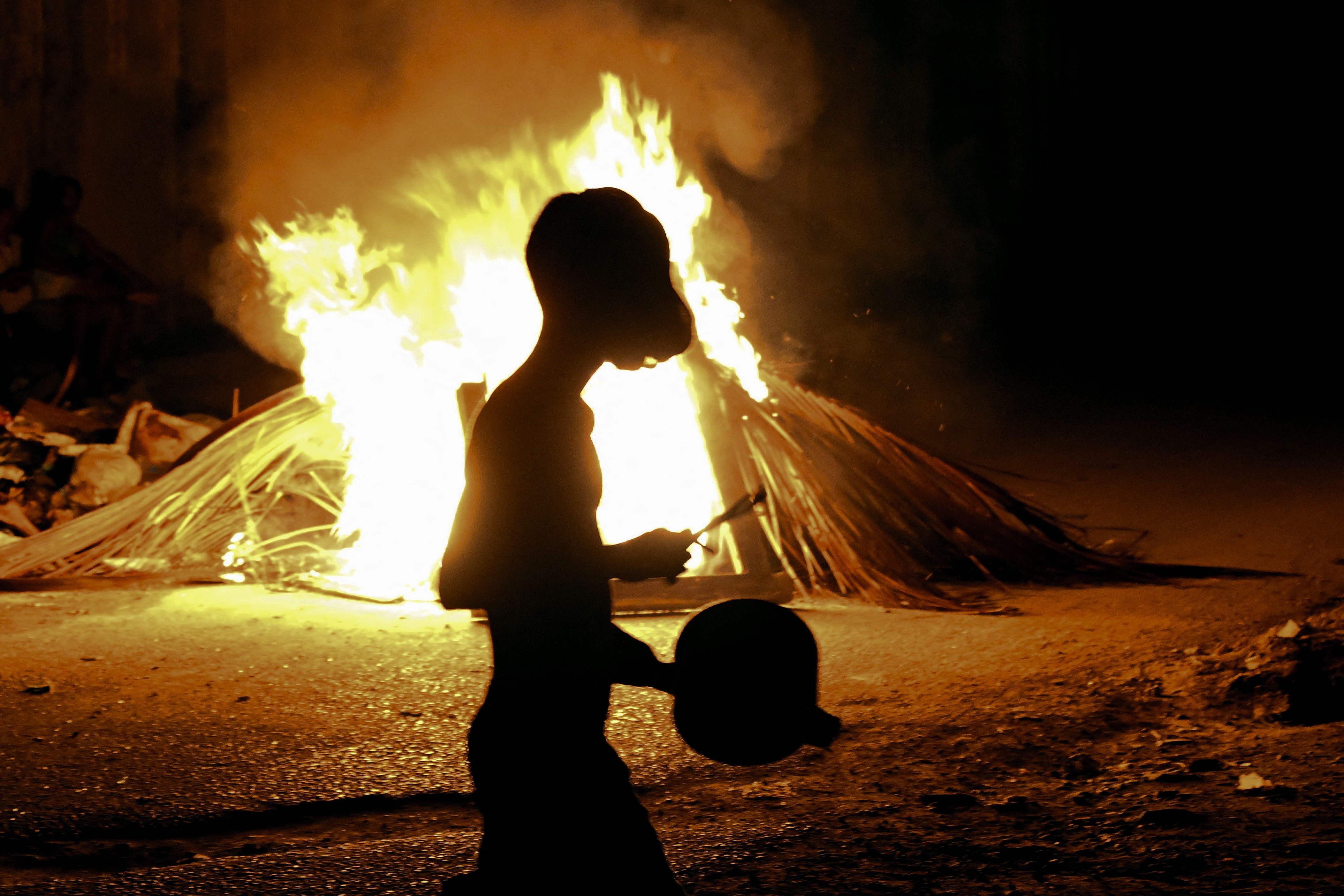 A man bangs a pot while walking past a fire set during a protest.