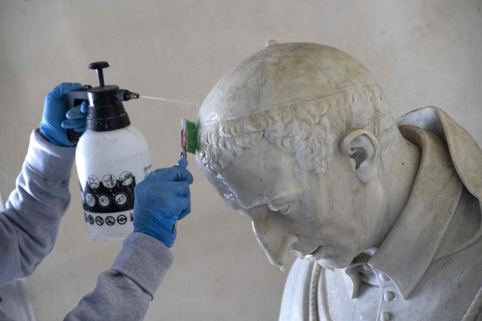 A worker sprays cleaner on the head of a statue, scrubbing with a brush.