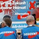 Two AfD supporters wearing the party's vests sit in front of an AfD sign.