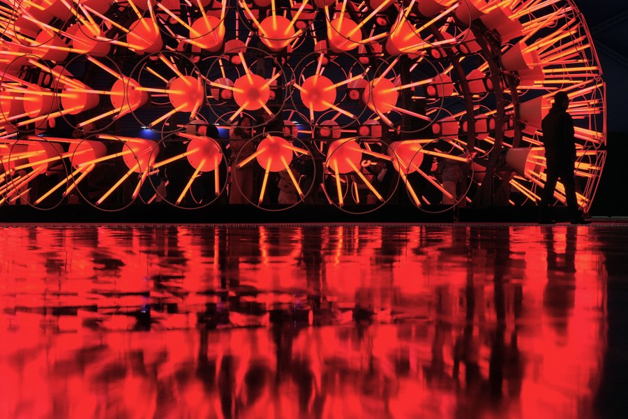 A person stands next to a large illuminated art structure that seems to be reflected in water.