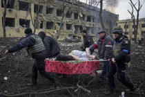 Five men carry a pregnant woman on a stretcher. They are surrounded by destroyed buildings and debris.