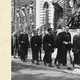 A black and white photograph of male Harvard students from the 1950s in graduation robes processing past an ivy-lined building and the John Harvard statue