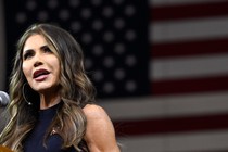Kristi Noem speaking at a podium in front of a giant American flag