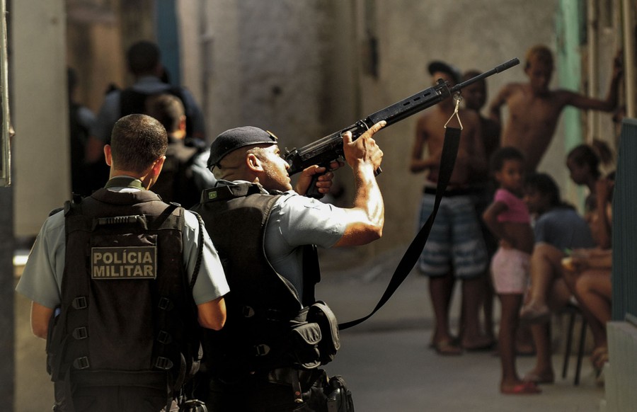 Several police officers walk past children in an alley, as one officer holds up a rifle.
