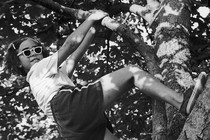 A black-and-white image of a girl smiling as she hangs from a tree branch, with one foot propped against the tree's trunk