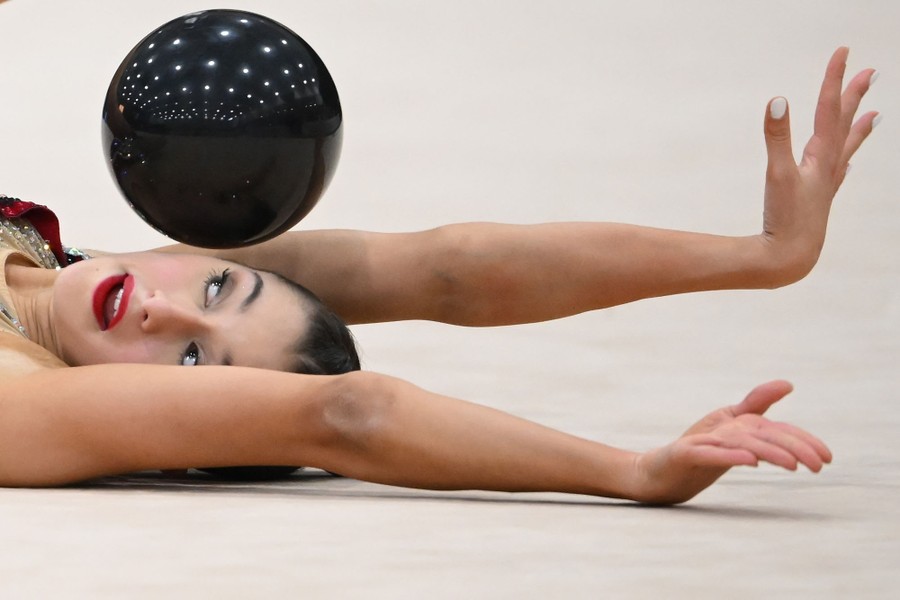 A gymnast lies on the floor, arms outstretched during a routine, with a small ball near their face.