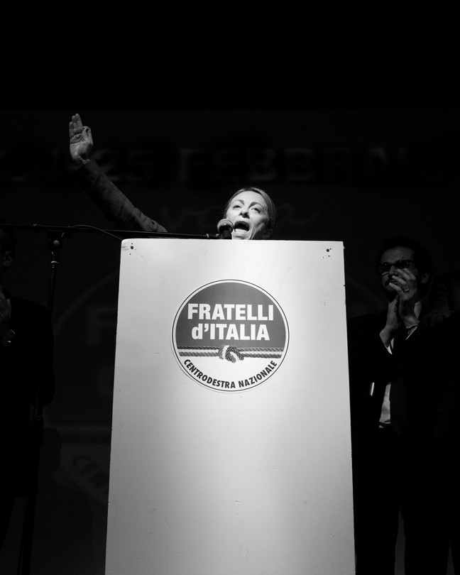 Black-and-white image of Meloni raising her arm in the air behind a podium as she talks into the microphone