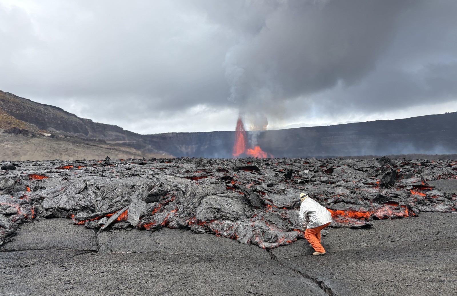 A person in protective gear leans close to an active lava flow, reaching out to gather a sample.