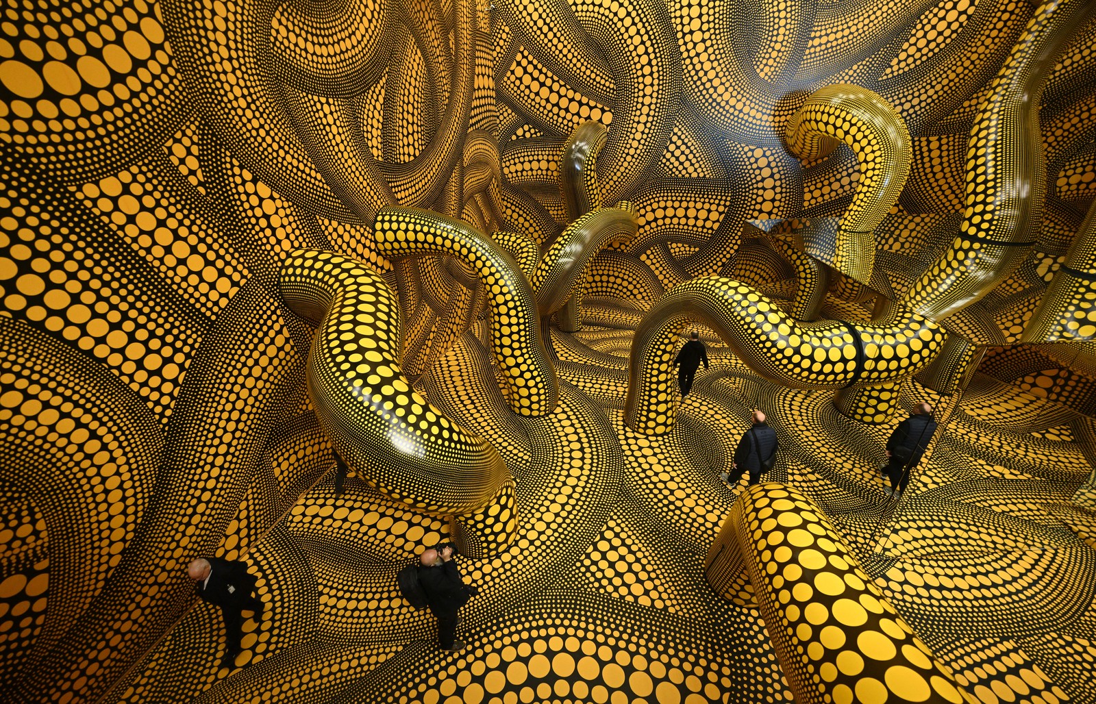 Visitors walk through a large room-sized art installation resembling dozens of twisting tubes decorated with yellow circles on black backgrounds.