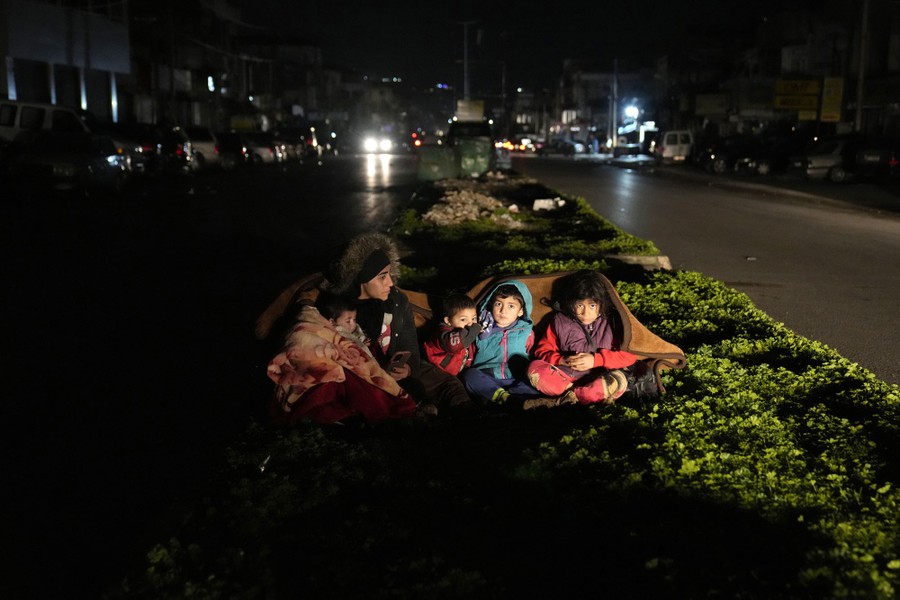 A woman and four children wrap themselves in blankets, sitting on a divider in the middle of a road at night.