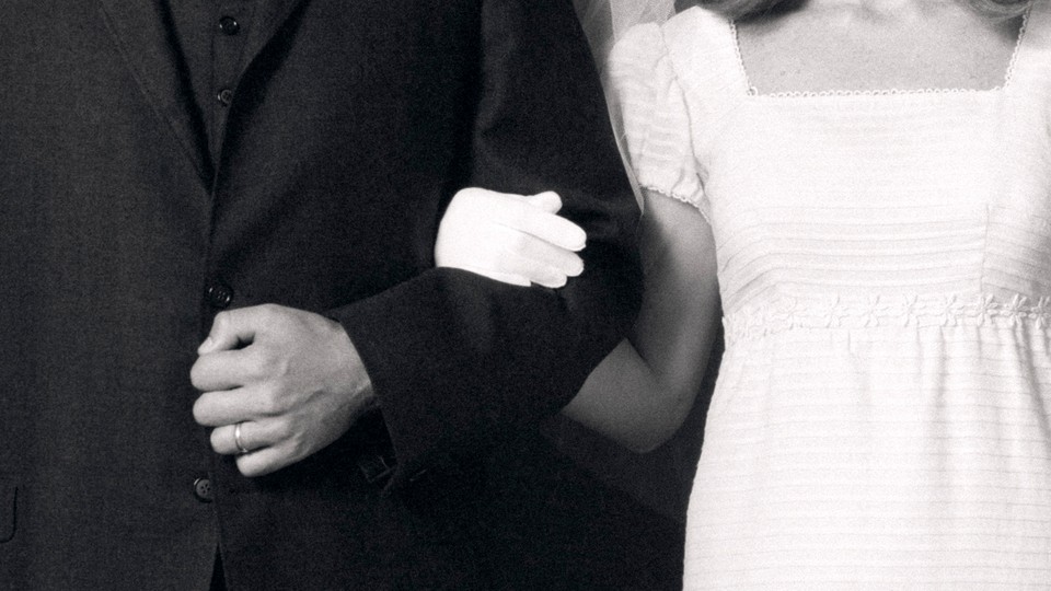 A black-and-white photo of man in a tux and a woman in a white dress, with arms locked