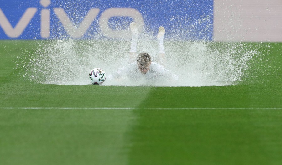 A soccer player slides facedown on soaked grass, sending up splashes of water.