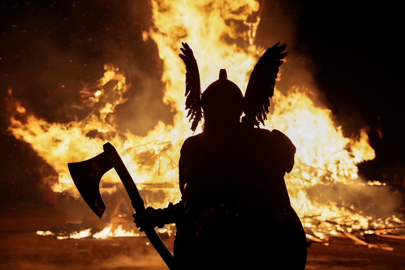 A person wearing a fanciful Viking costume, holding an axe, stands in front of a burning replica of a viking ship, seen in silhouette.