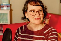 a portrait of the cartoonist and author Raina Telgemeier, wearing a red striped sweater and sitting in her home, in front of a bookshelf of her work.