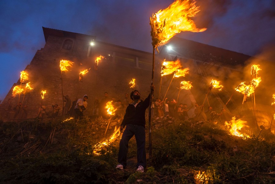 A group of people stand on a hillside, holding torches on long poles.