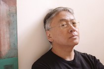 The author Kazuo Ishiguro leaning against a wall with two paintings visible