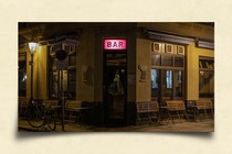 A bar at night, with an illuminated pink sign in the center and folded-up tables along its windows